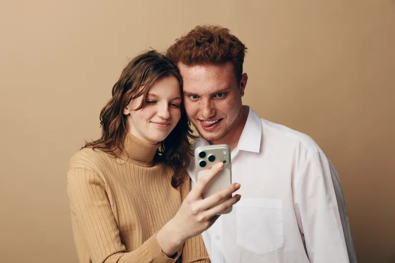 Young couple takes selfie with smartphone, smiling and enjoying moment together. Casual clothing and warm beige background create cozy and intimate atmosphere.