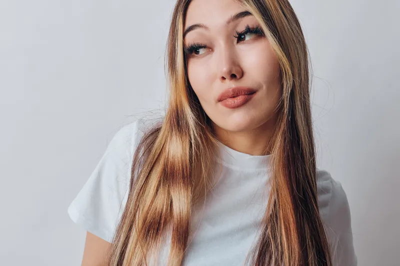 Woman with long hair wearing white t shirt looking aside with thoughtful expression on neutral background. Portrait of female model with natural makeup and casual style indoors.