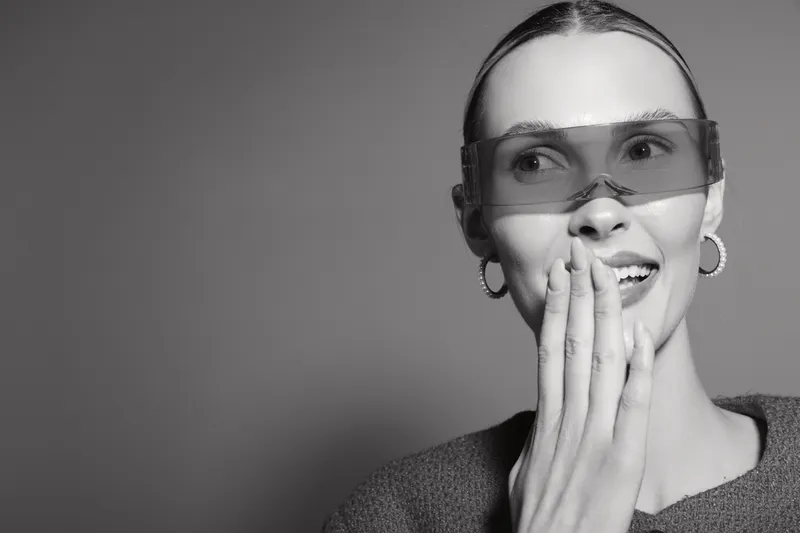 Young caucasian woman with light skin wearing transparent glasses covers mouth with hand, smiling and looking aside, isolated on grey background.