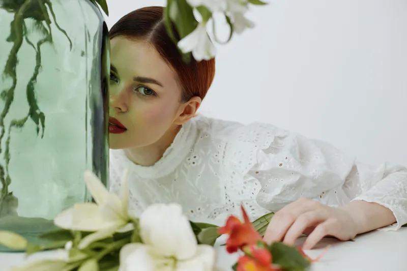 Young woman with red hair peeking from behind a glass vase, adorned in a white lace dress, surrounded by vibrant flowers, embodying beauty and freshness