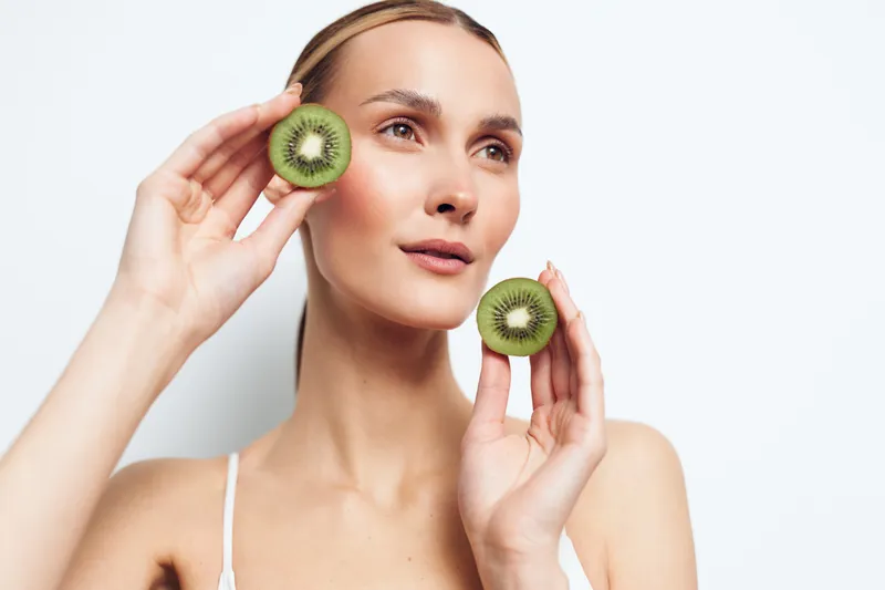 A young woman holds two halves of a fresh kiwi fruit near her face, looking away thoughtfully. The bright studio background emphasizes healthy lifestyle and natural beauty concepts.