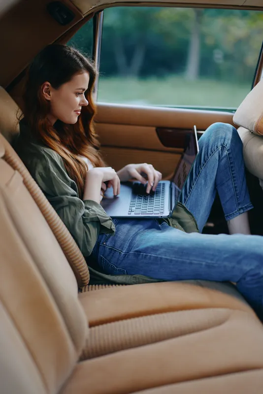 Young woman working on laptop in car, stylish casual outfit, focused expression, natural lighting, outdoor background, perfect for freelance lifestyle and inspiration.