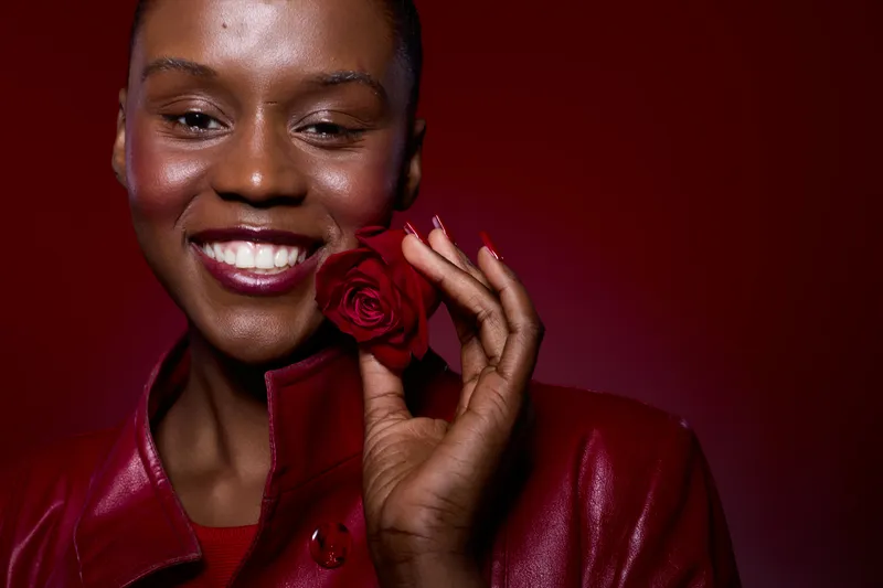 Close-up portrait of a smiling dark skinned African woman holding a red rose near her face. She wears a red leather jacket against a red background. Concept of beauty and confidence.