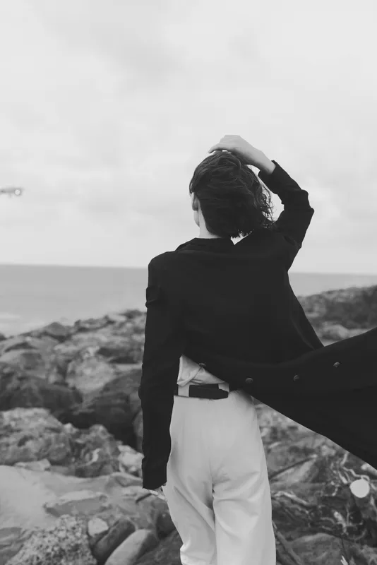 Cape and coat clad silhouette facing the sea on a rocky shore, wind lifting fabric in a dramatic outdoor scene with a moody horizon and stylish fashion mood