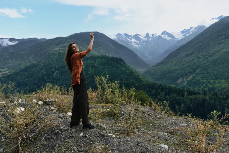 Woman standing on a rocky hillside in a scenic mountain landscape, raising her arm to take a photo, with trees and distant snow peaks.