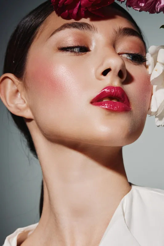 Close-up of a stylish young woman with glossy red lips and luminous skin. Creative fashion beauty portrait with flowers framing her face on a simple gray background.