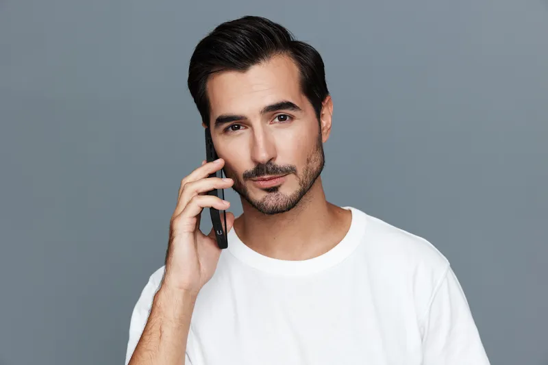 Handsome confident man talking on mobile phone, wearing casual white t-shirt, smiling, looking at camera, against plain solid grey background. Lifestyle concept.