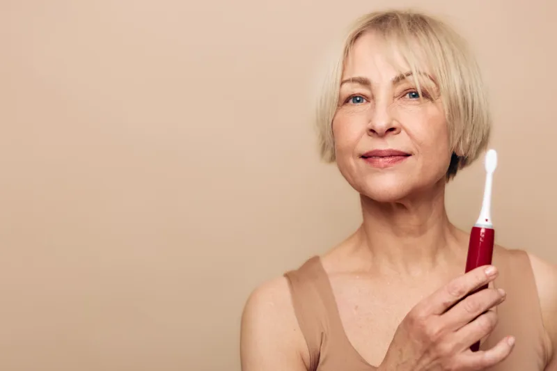 Mature woman with short blonde hair holds electric toothbrush smiling gently on beige background. Concept for dental care, oral hygiene, wellness, and healthy lifestyle at home.