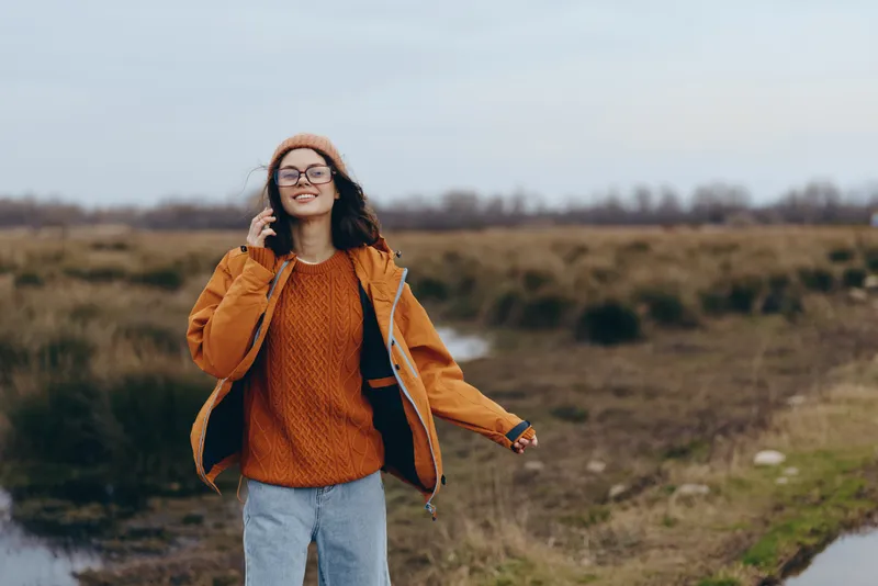Woman lifestyle outdoors, smiling in an open field, wearing orange sweater and jeans, beanie and glasses, relaxed pose by rural path, soft breeze, calm autumn mood morning light today