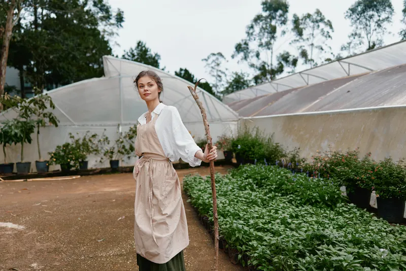 Young woman in a garden wearing a white shirt and apron, holding a stick among lush greenery, showcasing sustainable gardening and a serene atmosphere