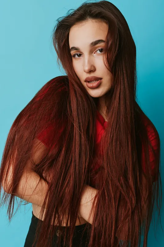 Young woman with long brown hair posing against blue background wearing red top. Beauty portrait with natural makeup and confident expression in studio lighting.