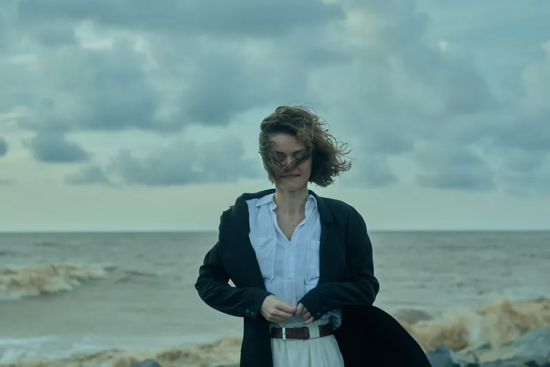 Business woman on a windy shoreline adjusts her coat as waves crash, projecting resilience, coastal mood and professional presence against a dramatic sea backdrop