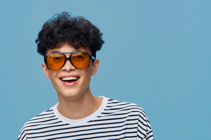 Smiling man wearing orange sunglasses and striped shirt, happy young adult with curly hair against light blue background, casual style, positive mood.