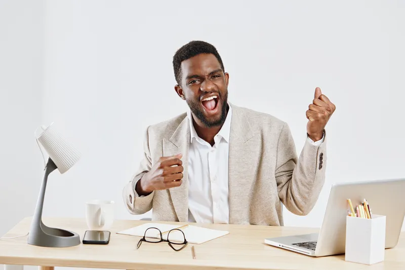 Confident young man in formal blazer celebrating success at modern office desk, showing excitement and joy, with laptop, glasses, and stationery, on clean background. Business achievement concept.