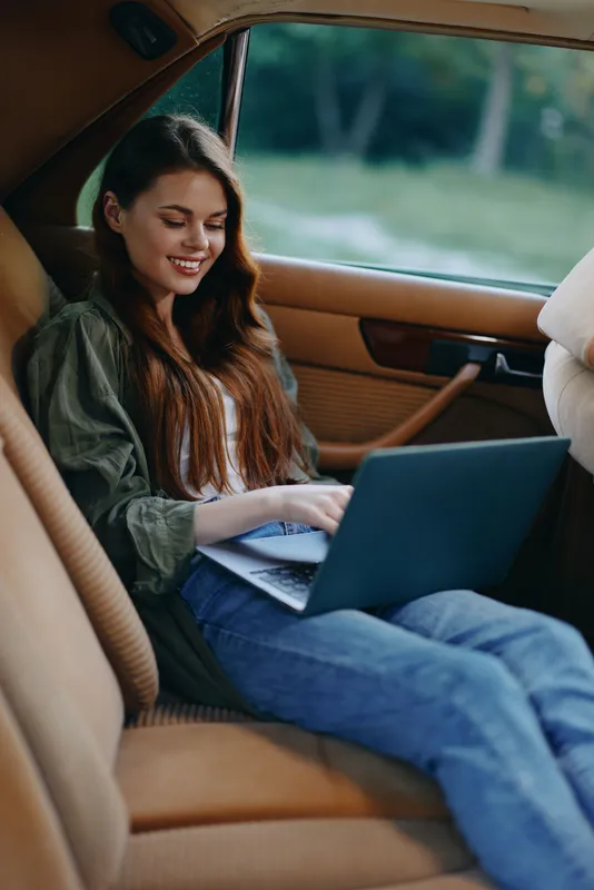 Young woman working on laptop in a car, dressed casually, smiling happily, showcasing productivity and comfort in a serene outdoor setting.
