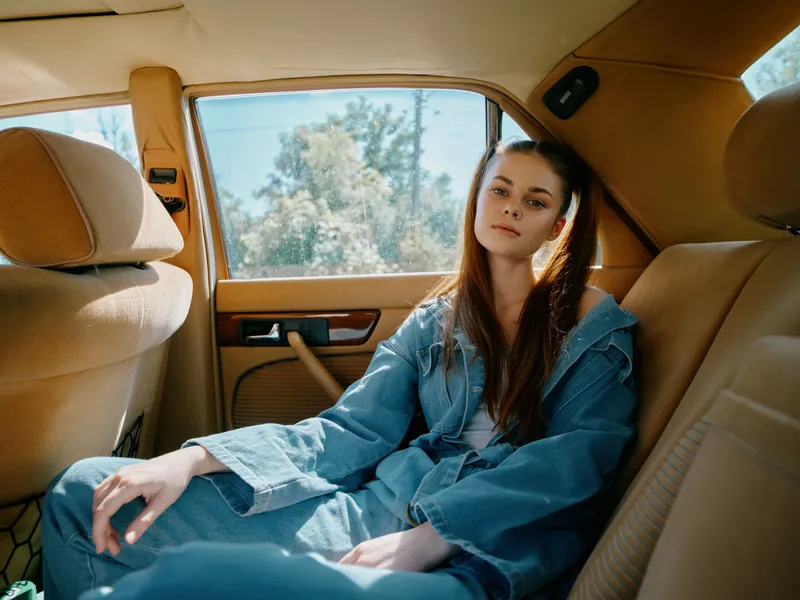 Young woman in denim outfit sitting in a retro car, showcasing a relaxed expression and natural sunlight, conveying a sense of freedom and nostalgia.
