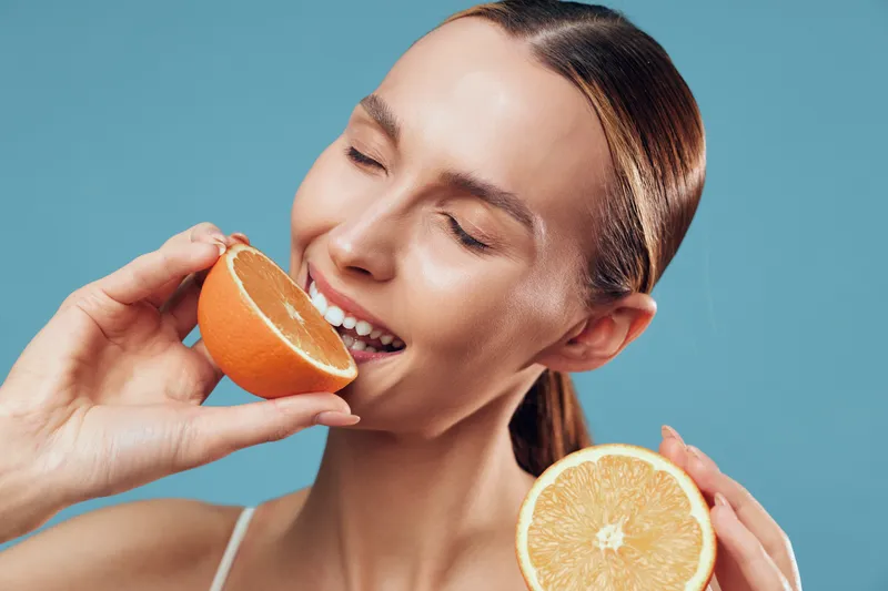 A young woman smiles with her eyes closed as she bites into fresh orange halves, vibrant juices catching the light against a bright blue background, conveying playful energy and healthy snacking.