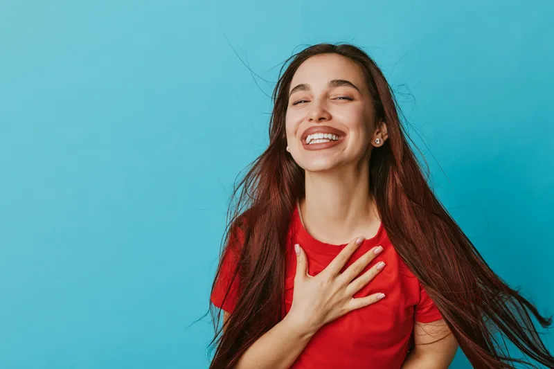 Young woman with long hair smiling with eyes closed and hand on chest wearing red shirt against blue background for happiness and gratitude concept.