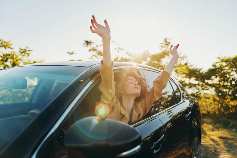 Woman leaning out of a car window with arms raised under bright sunlight, capturing a carefree lifestyle, candid moment, retro film color, warm glow, outdoor freedom.
