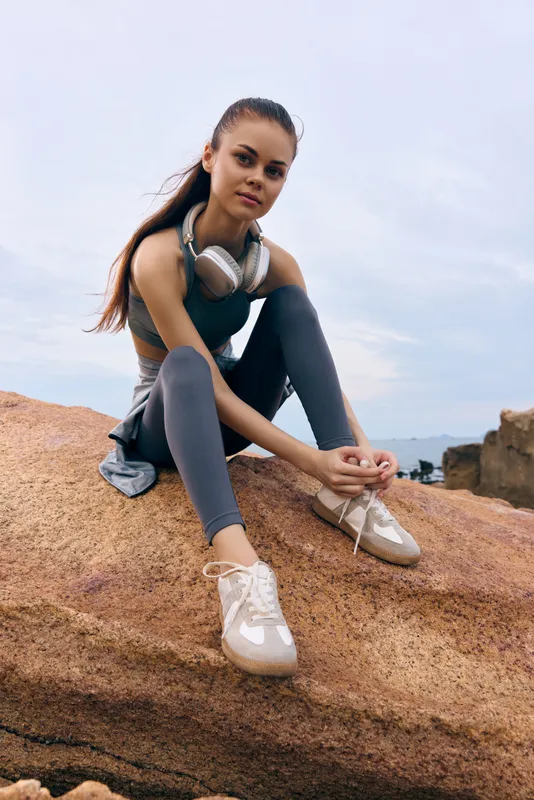 Young athletic woman relaxing on a rock in activewear with headphones, embodying a focus on fitness and tranquility in a natural setting with a serene sky