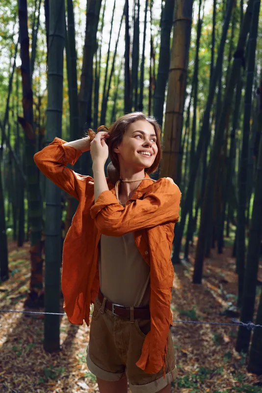 Woman smiling in a bamboo forest portrait, outdoor travel scene with orange jacket and shorts, sunlight filtering through trees, joyful nature moment and casual lifestyle expression