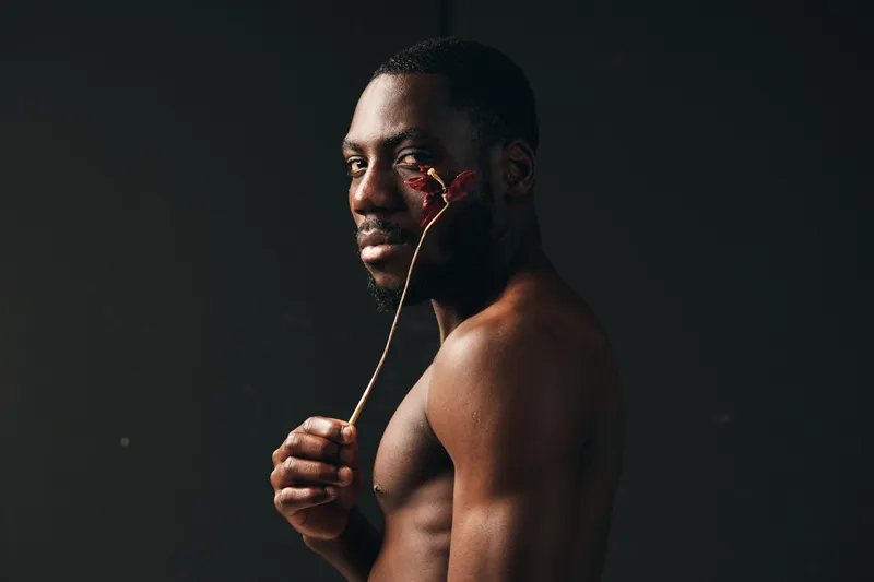 African man with dark skin holding dried flower close to face in creative conceptual visual style. Portrait with bare torso and serious expression on black background in studio lighting.