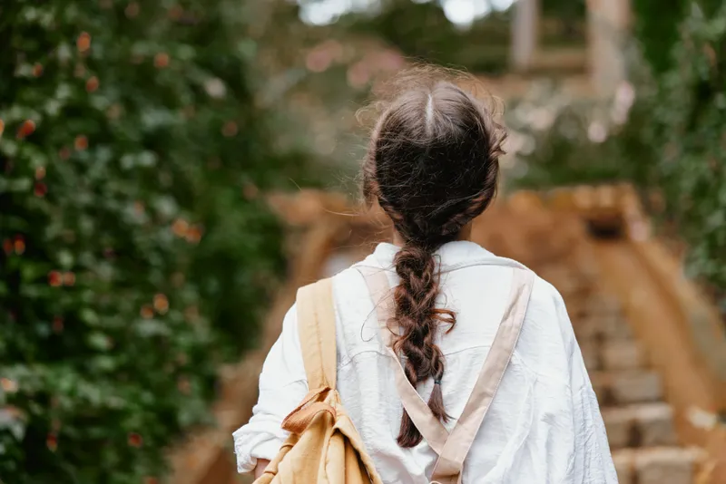 Young woman walking in nature with a braid, wearing a casual outfit and backpack, surrounded by greenery and tranquil scenery, evoking feelings of adventure and freedom