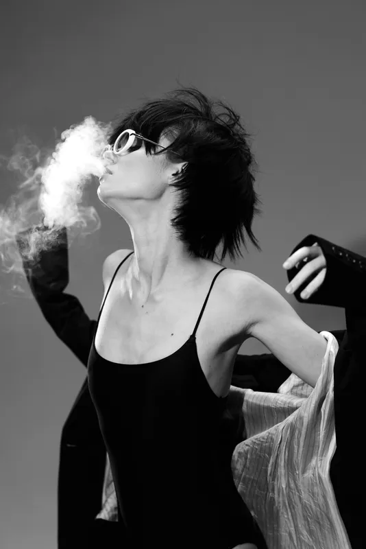 Black and white portrait of a young woman in sunglasses exhaling smoke with stylish outfit and dynamic pose.