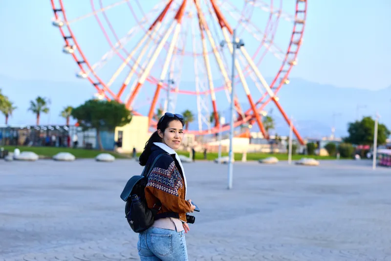 Young woman in casual jacket and jeans with backpack pauses near a towering ferris wheel at an amusement park, outdoor scene, exploring and posing for travel photography.