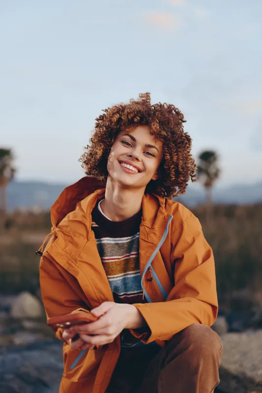 Woman lifestyle portrait outdoors, smiling with curly hair, wearing orange jacket, sitting on roadside path, warm sunset light, relaxed mood, countryside background, soft focus.