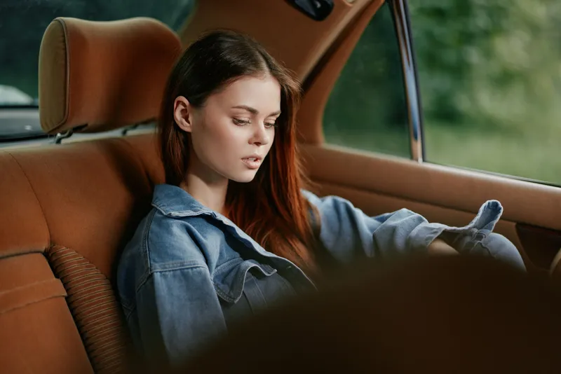 Young woman in denim jacket sitting in the backseat of a car, reflecting on life with a pensive expression, surrounded by nature.