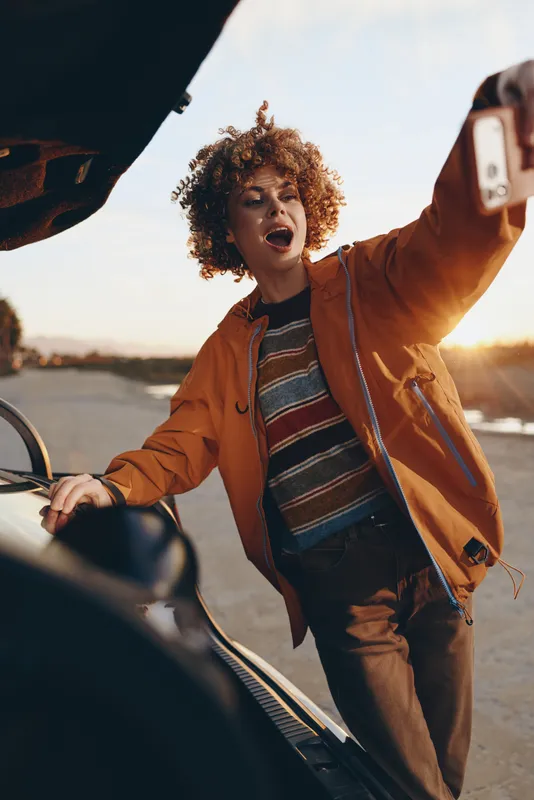 Woman lifestyle selfie with smartphone near car on road at sunset, curly hair outdoors, wearing jacket and scarf, joyful smiling expression, wind and warm daylight, closeup portrait