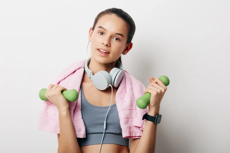 Woman fitness enthusiast holding light dumbbells with a towel draped over shoulders and headphones, showcasing a focused workout routine in a bright studio.