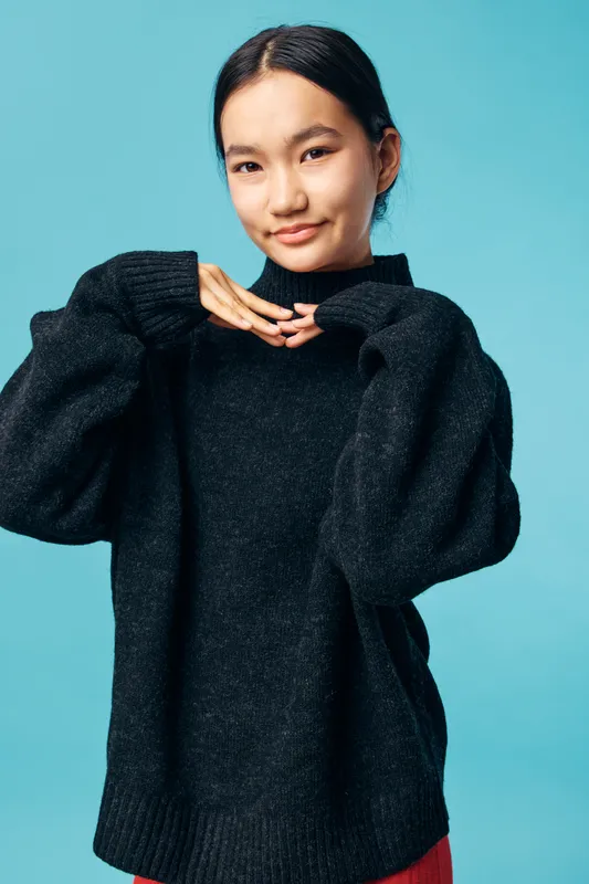 Young woman posing in black oversized sweater with hands near chin against light blue background in studio. Casual, comfortable, fashion, portrait of confident female model with smile.