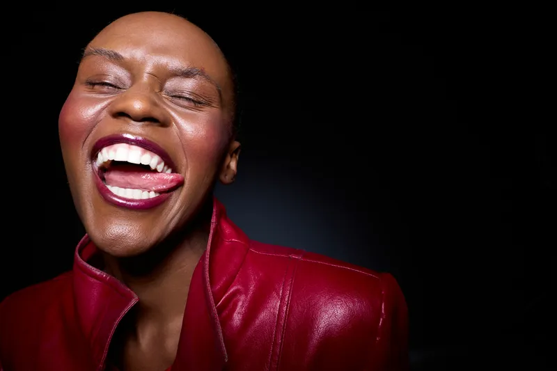 Close-up of a happy black woman with dark skin laughing joyfully wearing a red leather jacket, isolated on a black background. Concept of happiness, fashion, and natural beauty.