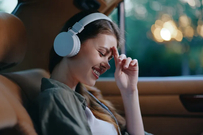 Young woman enjoying music with headphones inside a car, conveying happiness and relaxation during a sunset.