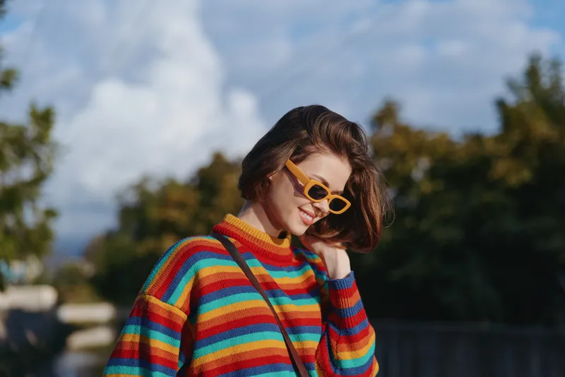 Young woman in rainbow sweater and yellow sunglasses smiling outdoors, short brown hair, casual crossbody bag, leaning head in sunny park scene for lifestyle fashion and travel vibe.