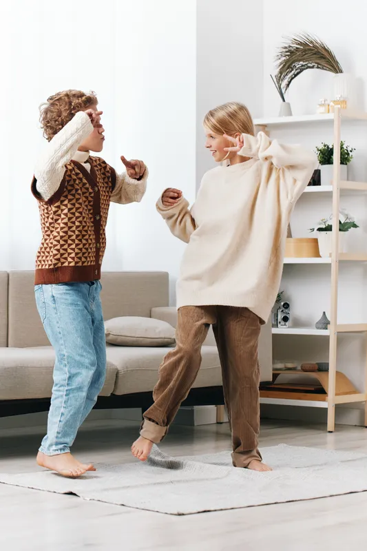 Two children dancing indoors in a bright living room, wearing cozy sweaters and jeans, capturing carefree energy, movement, and joyful friendship in a playful, wholesome scene.