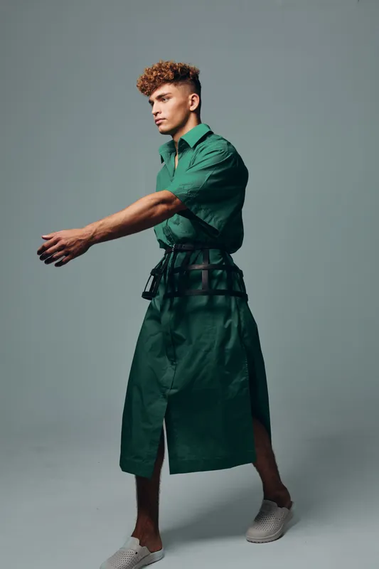 Young man with curly hair in a long green dress and white shoes posing on gray studio background with serious expression and outstretched arms showing fashion and style.
