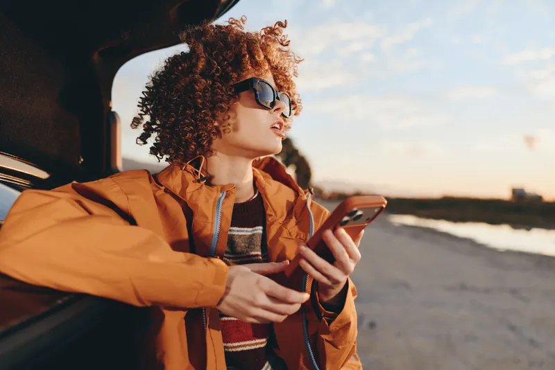 Woman lifestyle, curly hair, by car window at seaside sunset, holding smartphone, casual travel portrait, orange jacket, warm golden light