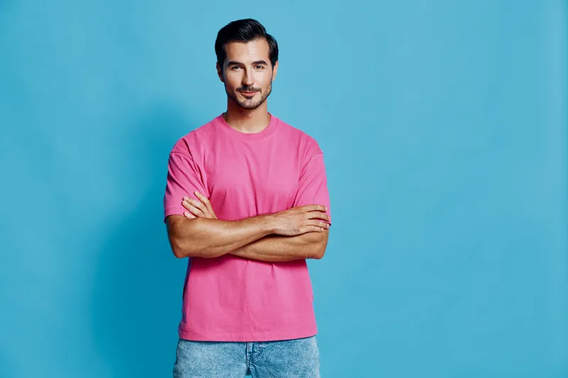 Smiling man in pink t-shirt poses confidently with arms crossed against a vibrant blue background, exuding a relaxed and approachable vibe. Fashion lifestyle concept.
