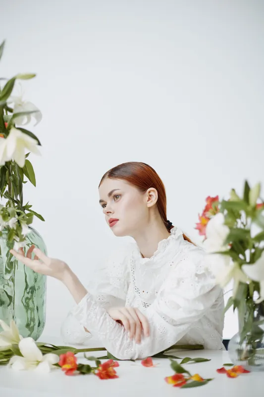 Elegant young woman in a white blouse, surrounded by colorful flowers, conveying calmness and beauty in a minimalistic setting