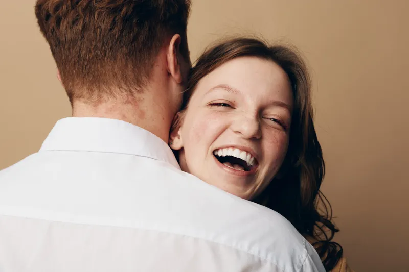 Happy young woman with curly hair laughs joyfully while hugging a man in white shirt. Close-up portrait on beige background. Concept of love, happiness, and relationship.