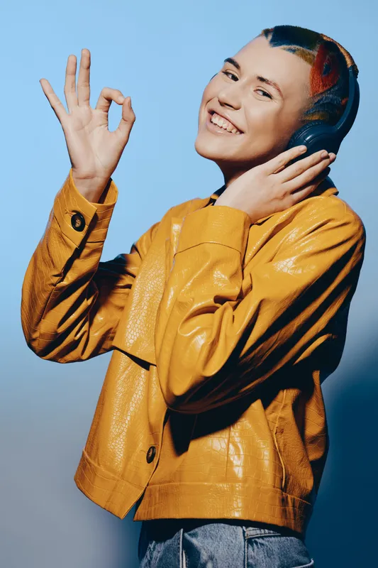 Young woman with headphones smiling happily, wearing a bright yellow jacket, showing okay hand gesture, colorful hairstyle, casual style, vibrant mood.
