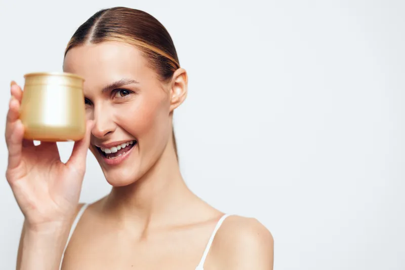 Cheerful woman with a gold cup held near her lips, bright smile, studio portrait. Beauty wellness spa relaxation concept with clean skincare and minimal soft white background.