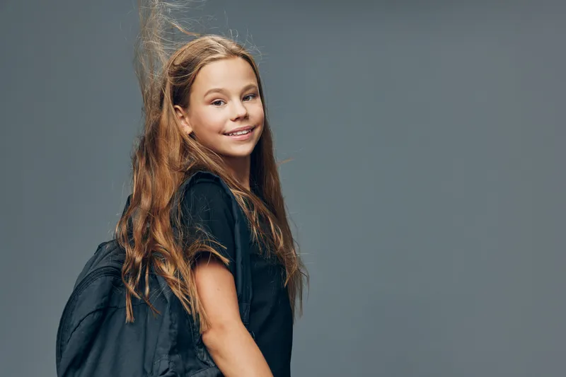 Girl student standing with backpack and smiling, young female child posing for camera with casual black clothing on neutral background, education and youth concept.