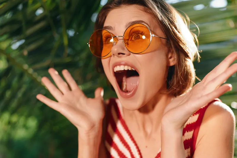 A joyful woman wearing round orange sunglasses is outdoors, expressing excitement with a wide smile and open hands, creating a bright, energetic summer vibe.