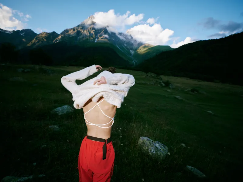 Athlete in red pants twirls in an open back sweater against a mountain landscape, showcasing freedom of movement and bold fashion in a wild green valley.