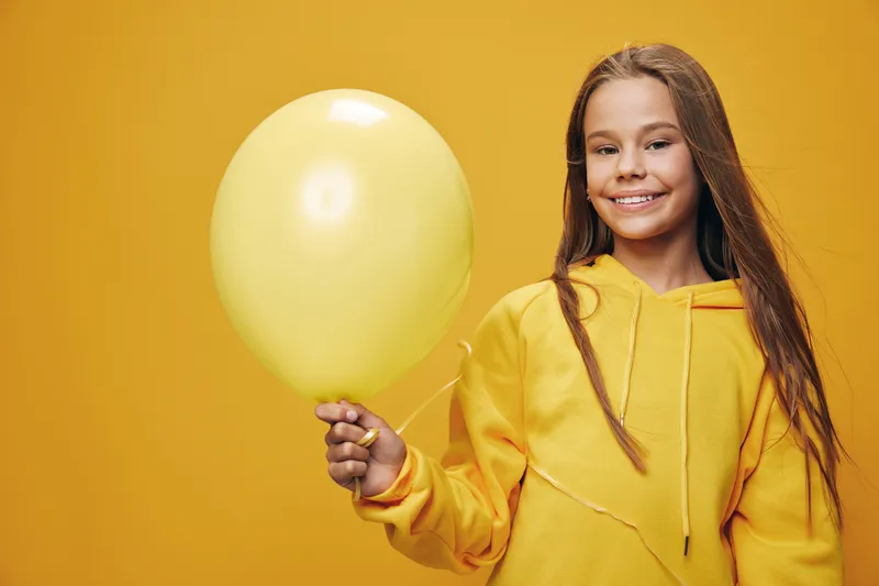 Girl smiling and holding yellow balloon in hand wearing yellow hoodie standing against yellow background. Child portrait with long hair and cheerful expression indoors.