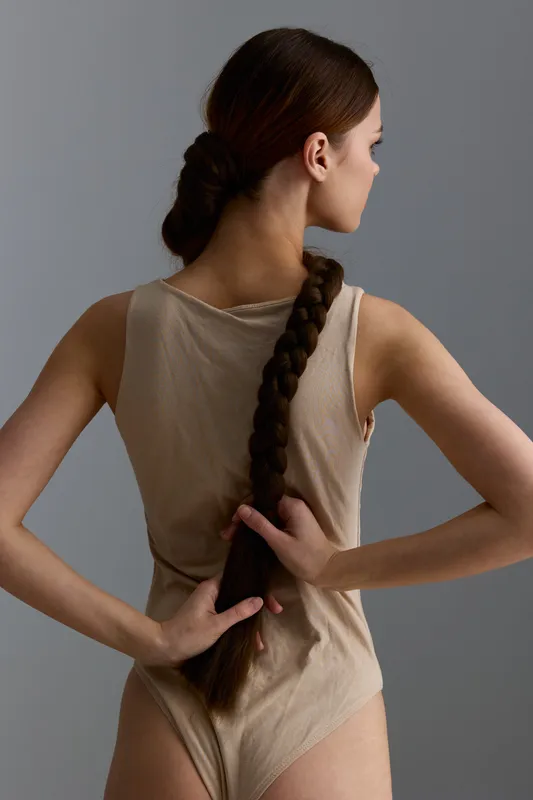 woman with long braid hairstyle wearing beige bodysuit poses with her back to the camera against gray background in minimalist studio setting